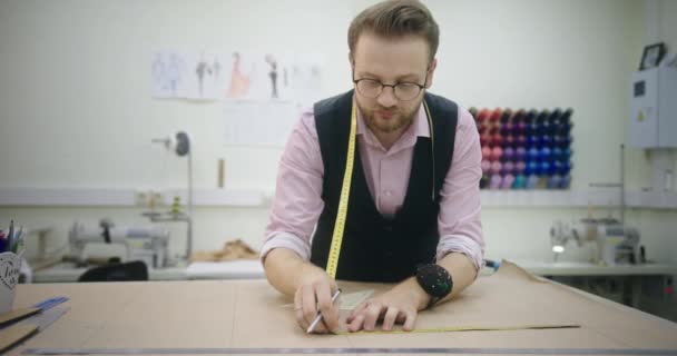 Un homme avec des lunettes un jeune tailleur dessine des mesures sur les flans pour la future robe du produit en tissu debout dans son studio propriétaire de petite entreprise — Video