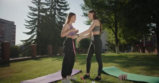 Zwei Mädchen messen die Parameter des anderen mit Hilfe eines Maßbands Freundinnen lächelnd im Sommer nach dem Training auf dem grünen Rasen stehen — Stockvideo