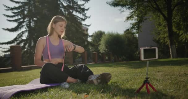 Menina bonita jovem sentado em um tapete depois de um treino de esportes olha para sua pulseira de fitness bebe água sorrindo sentado ao ar livre fora — Vídeo de Stock