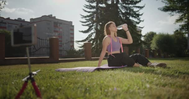 Młoda piękna dziewczyna siedząca na ulicy na macie na zielonej trawie po aktywnym treningu odpoczywająca woda pitna ubrana w strój sportowy — Wideo stockowe