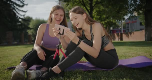 Dvě přítelkyně sedí spolu na zelené trávě pózování sledování videa a fotografie na smartphone smát po tréninku v letních sportů — Stock video