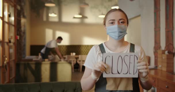 Comida interior cafetería cuarentena chico camarero da la vuelta sillas chica de pie mira a la cámara en máscara y mitones mantiene el plato cerrado — Vídeo de stock