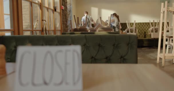 Un letrero está cerrado en la mesa de un restaurante cerrando un restaurante debido a una pandemia de virus dos camareros entregan sillas en una habitación soleada — Vídeos de Stock