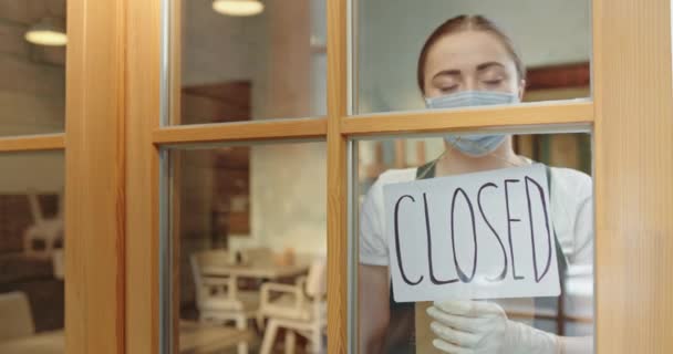 Flickan servitrisen står i en mask anställd av restaurangen vänder över en tallrik stängd på grund av en pandemisk karantän sorgligt besviken blickar på gatan — Stockvideo