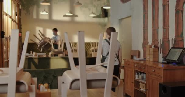 Dos camareros un tipo y una chica con máscaras entregan las sillas cerrando la institución durante el período de cuarentena pandemia de crisis — Vídeo de stock