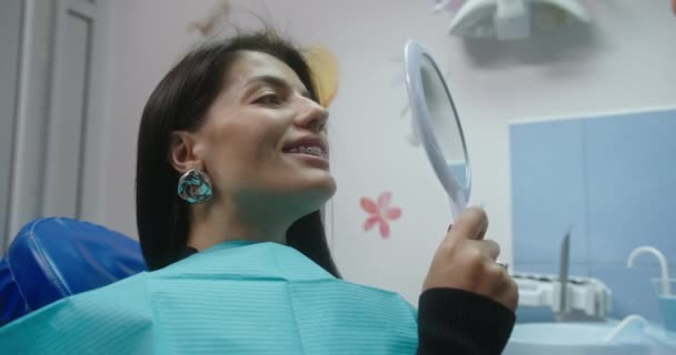 Un paziente con l'apparecchio si guarda attentamente allo specchio dopo aver installato l'apparecchio seduto su una sedia dentale in una clinica dentistica — Video Stock