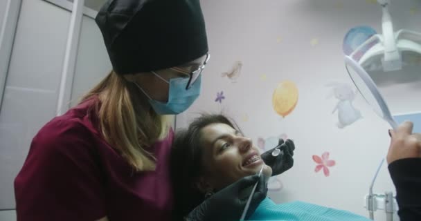 Прием пациента к женщине-ортодонту пациентке-стоматологу лежащей в стоматологическом кресле и смотрящей на себя с брекетами в зеркале — стоковое видео