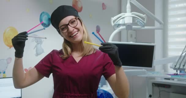 Демонстрация лигатур цветных резиновых лент для брекетов профессиональный стоматолог женщина-ортодонт, стоящая в стоматологическом кабинете улыбаясь — стоковое видео