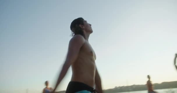 在夏天的日落时分，一个在海滨打沙滩排球的年轻活跃的小伙子和一群朋友一起发球 — 图库视频影像