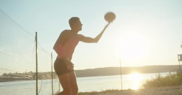 Actieve sportieve jonge man dient om bal stuiteren bij zonsondergang spelen met een team van vrienden in de zomer beachvolleybal aan de waterkant — Stockvideo