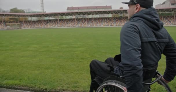 Ein Mann mit nicht funktionierenden Beinen, der im Rollstuhl im Stadion sitzt, blickt nach einer Verletzung traurig auf das Spielfeld. — Stockvideo