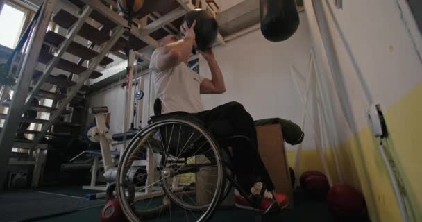 Entrenamiento en el gimnasio ex atleta hombre con las piernas defectuosas sentado en una silla de ruedas lanza la pelota entrena sus manos dentro — Vídeo de stock