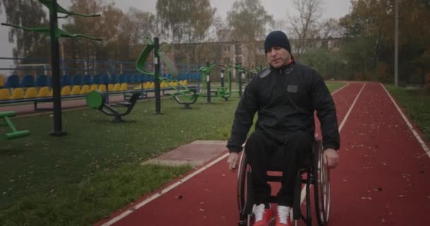 Voormalig atleet na blessure man met defecte benen, slopende rolstoel rijden op de straat van het stadion buiten, gekleed in sportkleding — Stockvideo