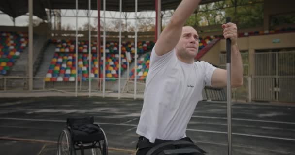 Kraftvoller Mann im Kugelstoßen mit defekten Beinen Rollstuhlfahrer führt Armwurfübung sitzend auf einer speziellen Halterung im Stadion aus — Stockvideo
