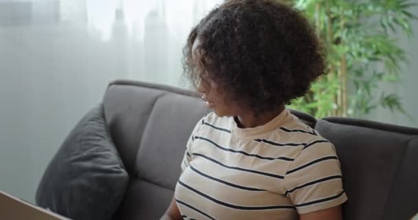 Freelancer afro girl trabajando en línea en casa sentado en el sofá mirando los gráficos de papel y trabajando mirando a la computadora portátil — Vídeos de Stock