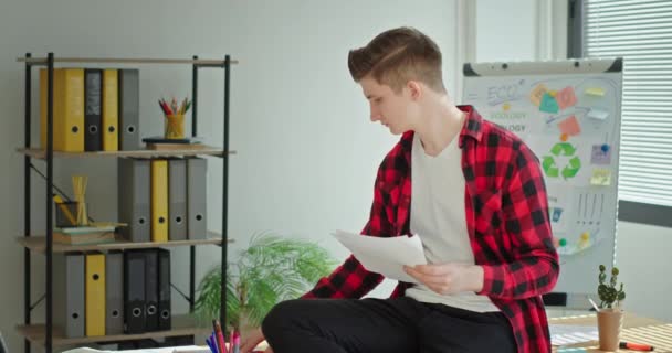 Junger Mann sitzt an einem Schreibtisch im Büro und schreibt Dokumente, während er in die Kamera lächelt und die Umwelt schont — Stockvideo