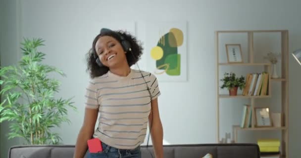 Ragazza afroamericana dalla pelle scura con i capelli ricci divertente ballare attivamente saltando in cuffia alla musica sorridente felice positivo a casa — Video Stock