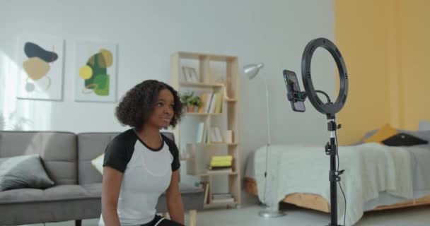 Mörkhyade afroamerikanska flicka register online klasser på telefonen upprätthåller en sport blogg leenden vinka hej och visar fitness övningar hemma på en matta — Stockvideo