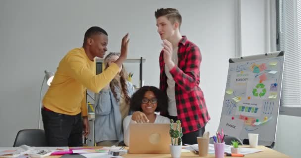 Egy csapat fiatal multiraciális dolgozik az irodában, a laptop előtt állnak és örvendeznek a boldog jó híreknek, és öt sikeres környezetvédelmi projektet adnak. — Stock videók