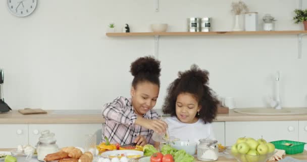 Zwei afroamerikanische Babymädchen mit lockigem Haar kochen zusammen und lachen positiv und machen Salat, der zu Hause in der Küche am Tisch sitzt. — Stockvideo