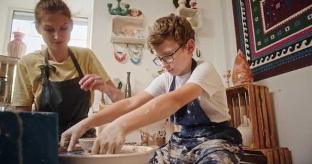 Mistr hrnčířská třída žena učí mladého chlapce pomáhá mu odstranit talíř s pomocí nástroje sedí za hrnčířským kolem — Stock video