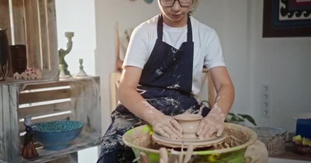 Młody nastolatek chłopiec siedzi garncarze koło patrząc w aparat uśmiechnięty zaangażowany w ceramiki tworzy gliniany produkt siedzi w kreatywnym studio — Wideo stockowe
