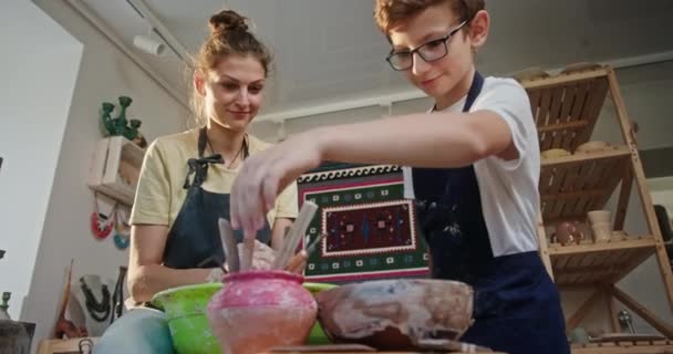 Pottery master class žena učí mladého chlapce zvážit nástroj pro kreativitu pozitivně usměvavý sedí za hrnčířským kolem — Stock video