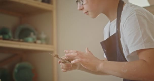 Giovane ragazzo adolescente crea un quadro su un piatto di argilla disegna uno strumento speciale è impegnato in ceramica seduto in una ruota vasai — Video Stock