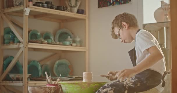 Молодий хлопчик-підліток, який займається гончарством, сидить у гончарному колесі, створює малюнок на гончарній картині спеціальний інструмент — стокове відео