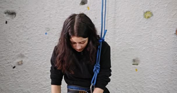 Pigen kigger ind i kameraet smilende positivt, at klatrer bliver klar til at klatre på en kunstig væg kontrollerer forsikringsforbindelserne med et reb. – Stock-video