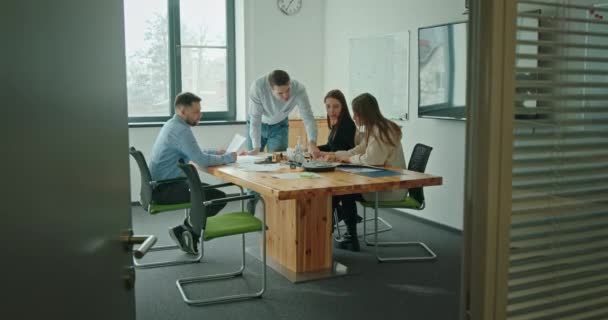Un equipo de jóvenes que trabajan juntos en una empresa en una oficina moderna sentados en una mesa de negociaciones discutiendo un nuevo proyecto un hombre dibuja un gráfico en una pizarra — Vídeo de stock