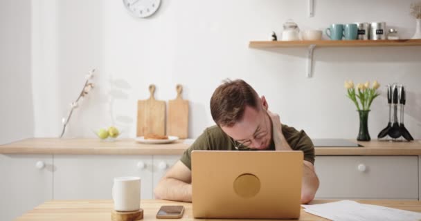 Tired man works on laptop massaging temples in kitchen — Stock Video