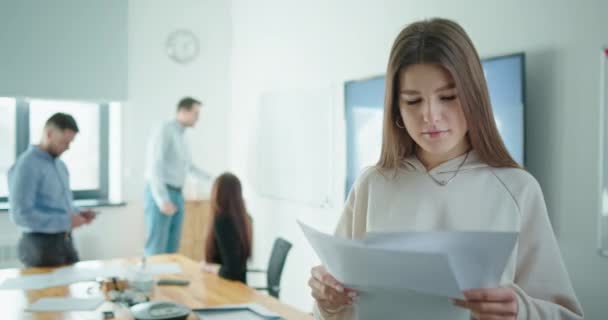 Junges Mädchen, das Papierdokumente in Händen hält, blickt emotional in die Kamera Team junger Menschen, die gemeinsam in modernem Büro arbeiten — Stockvideo