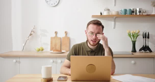 El hombre que duda con los cristales cansado del trabajo en el portátil en la cocina — Vídeos de Stock