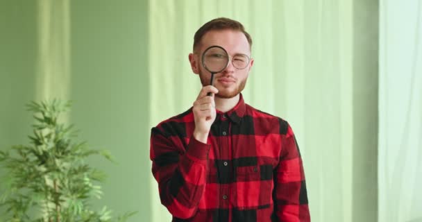 Divertido joven en gafas y una camisa a cuadros mirando a la cámara a través de una lupa investigador científico lupa de pie en el interior divertido video — Vídeo de stock