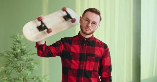 Jovem adolescente bonito olha para a câmera em pé dentro de casa pega um skate patins, endireita o cabelo — Vídeo de Stock