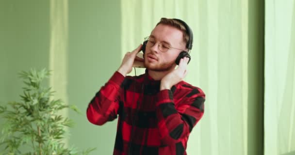 Chico bailando en grandes auriculares con teléfono bailando movimientos activos joven de pie en el interior mostrando emociones positivas sonriendo — Vídeo de stock