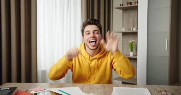 Joyful man in hoodie makes faces showing tongue at table — Stock Video