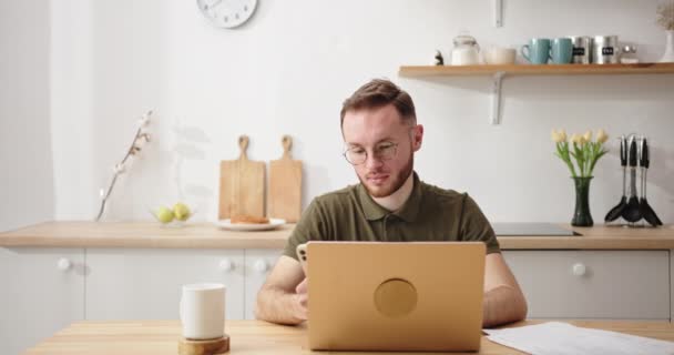 Νεαρός τύπος που λαμβάνει sms στο τηλέφωνο χαμογελώντας χαρούμενος κοιτάζοντας το τηλέφωνο κάθεται στο σπίτι στην κουζίνα στο τραπέζι εργασίας για laptop — Αρχείο Βίντεο