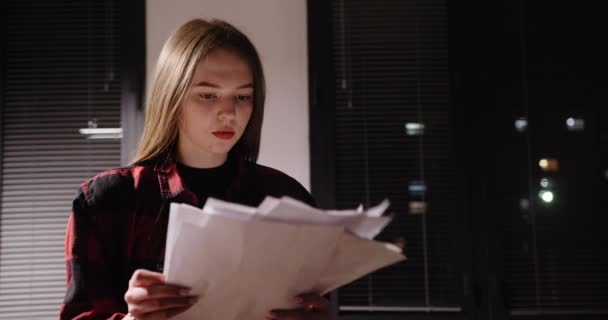 Νεαρό κορίτσι που εργάζεται τη νύχτα στο γραφείο κουρασμένος ψάχνει για χαρτί κοιτάζοντας μέσα από έγγραφα επαγγελματικά κοιτάζοντας γραφήματα που στέκονται στο παράθυρο μιας νυχτερινής πόλης — Αρχείο Βίντεο
