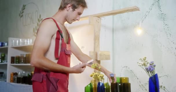 Un joven crea una composición toma fotos en el teléfono de sus jarrones de vidrio de procesamiento de cristalería con flores propias en su taller en ropa especial — Vídeo de stock