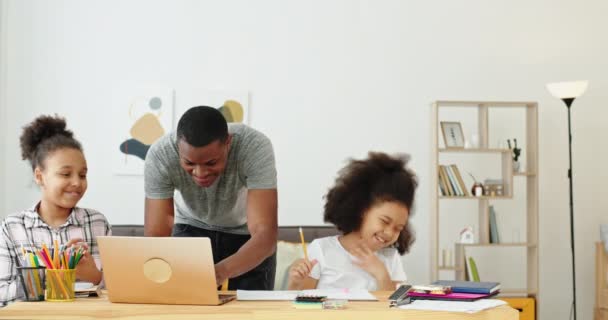 Nero afroamericano padre di famiglia insegna lezioni a casa con le sue figlie sedute a tavola e laptop imparare on-line divertimento sorridente emozioni positive — Video Stock