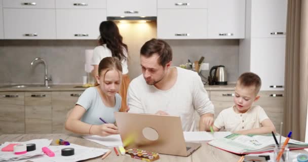 Vater sitzt mit Tochter und Sohn am Tisch und macht Hausaufgaben am Laptop, hat keine Zeit müde Kinder fordern Aufmerksamkeit Mama arbeitet im Hintergrund in der Küche in einer hellen Wohnung — Stockvideo