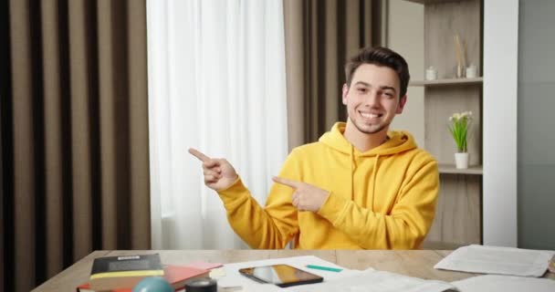 Demostración mostrando comercial disparo joven chico sonriendo señalando los dedos un lado sentado en casa en la mesa demostrando noticias de compras — Vídeo de stock