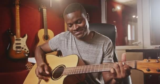Un tânăr muzician afro-american de culoare cântă activ la chitară, ținându-și degetele frumos, stând într-un studio de înregistrări atmosferice, pasionat de hobby-ul său, zâmbind, uitându-se la... — Videoclip de stoc