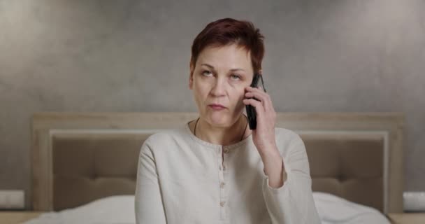 Серьезная раздраженная женщина, разговаривающая по телефону сердито отвечает закатывая глаза неприятный телефонный разговор откладывает телефон в сторону, сидя дома и в постели — стоковое видео
