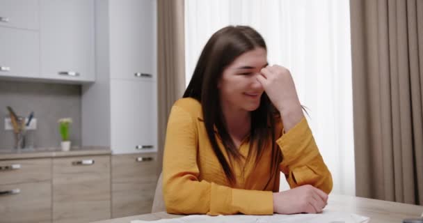 Schöne junge Frau, die an einem Tisch in einer hellen Wohnung sitzt, wendet sich beschämt glücklich lächelnd von positiven Emotionen ab — Stockvideo