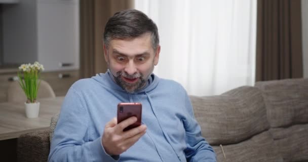 Starszy mężczyzna siedzący w domu na kanapie raduje się krzykami z radości zwycięstwa patrzy na telefon zdejmuje okulary raduje się uśmiechem z tego co zobaczył — Wideo stockowe