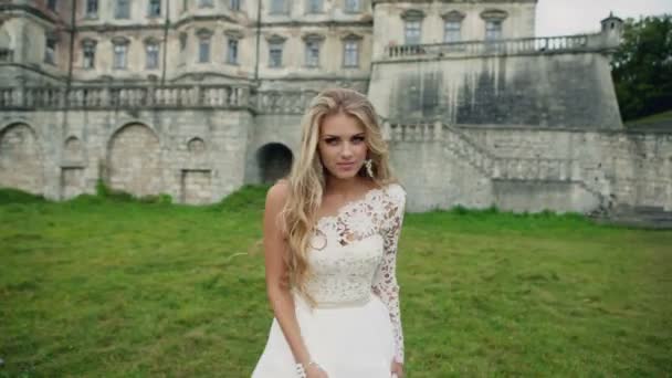 Giovane bella ragazza con lunghi capelli dorati in un abito bianco sorridente alla fotocamera e dinamicamente in movimento sullo sfondo di un vecchio castello. All'aperto — Video Stock