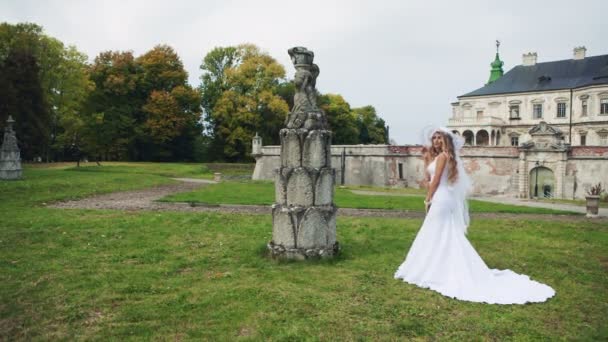 Дівчина в білій весільній сукні біля замку на зеленій траві дивиться на камеру . — стокове відео